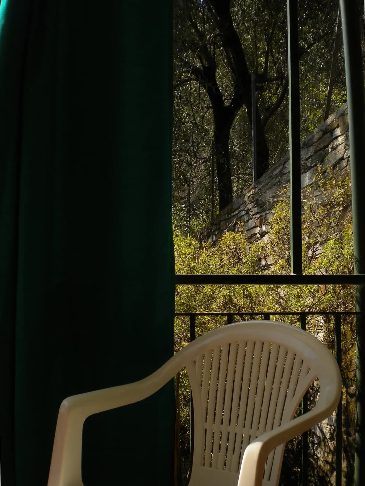 Una tenda verde scuro tirata su un balcone, accanto una sedia di plastica bianca e, oltre la ringhiera, un uliveto in pieno sole