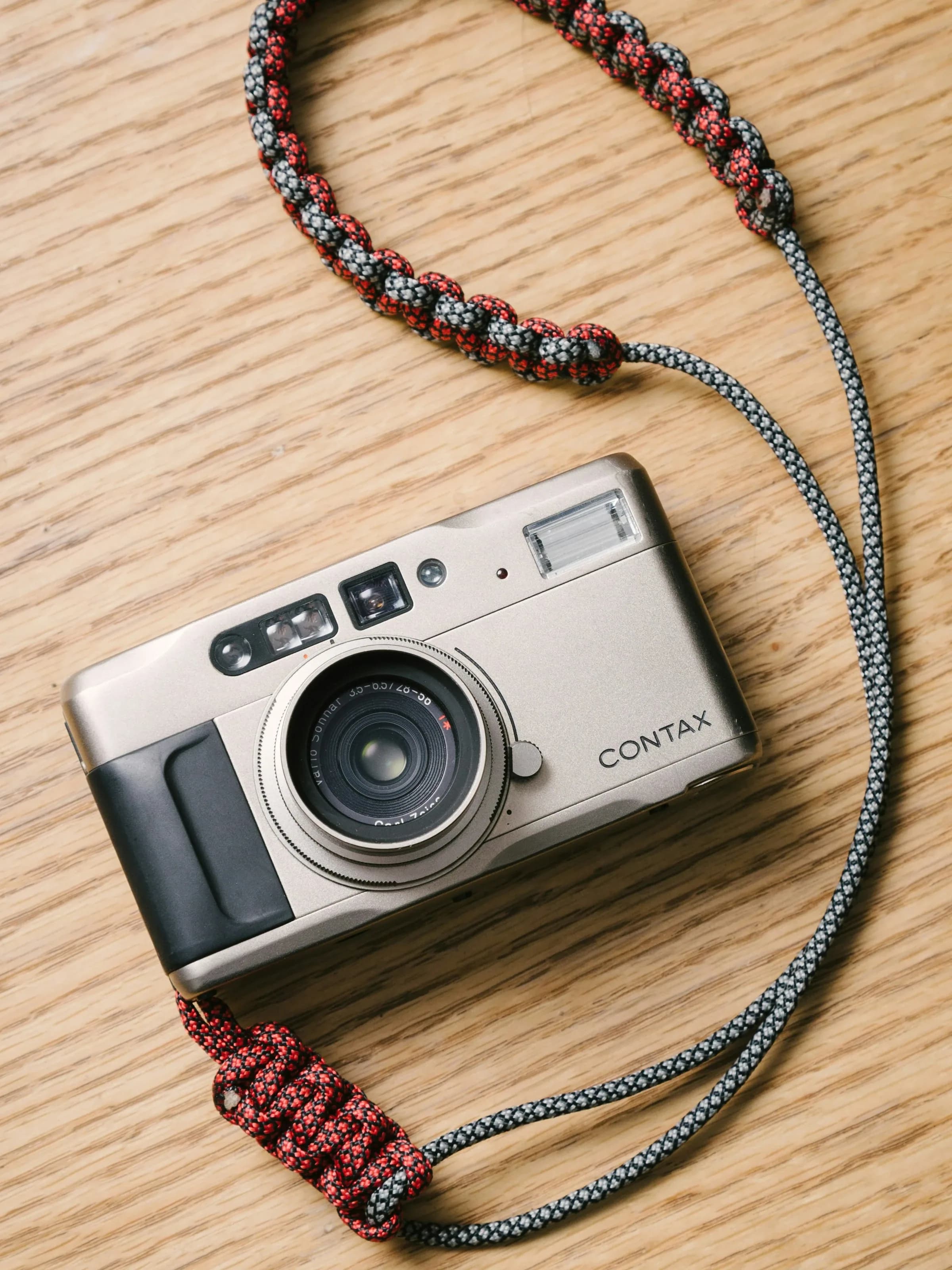 Una compatta Contax argentata su un tavolo di legno chiaro, con la cinghia intrecciata rossa e grigia