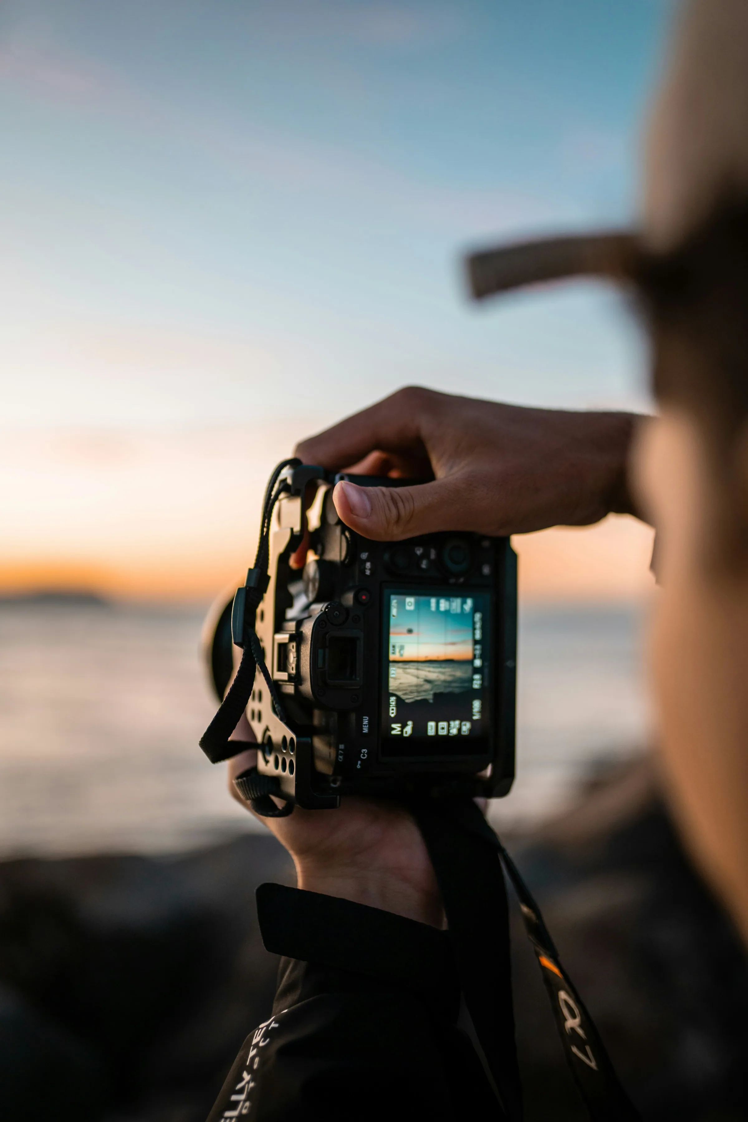 Una mano regge una fotocamera contro il mare al tramonto, l'inquadratura visibile sullo schermo posteriore