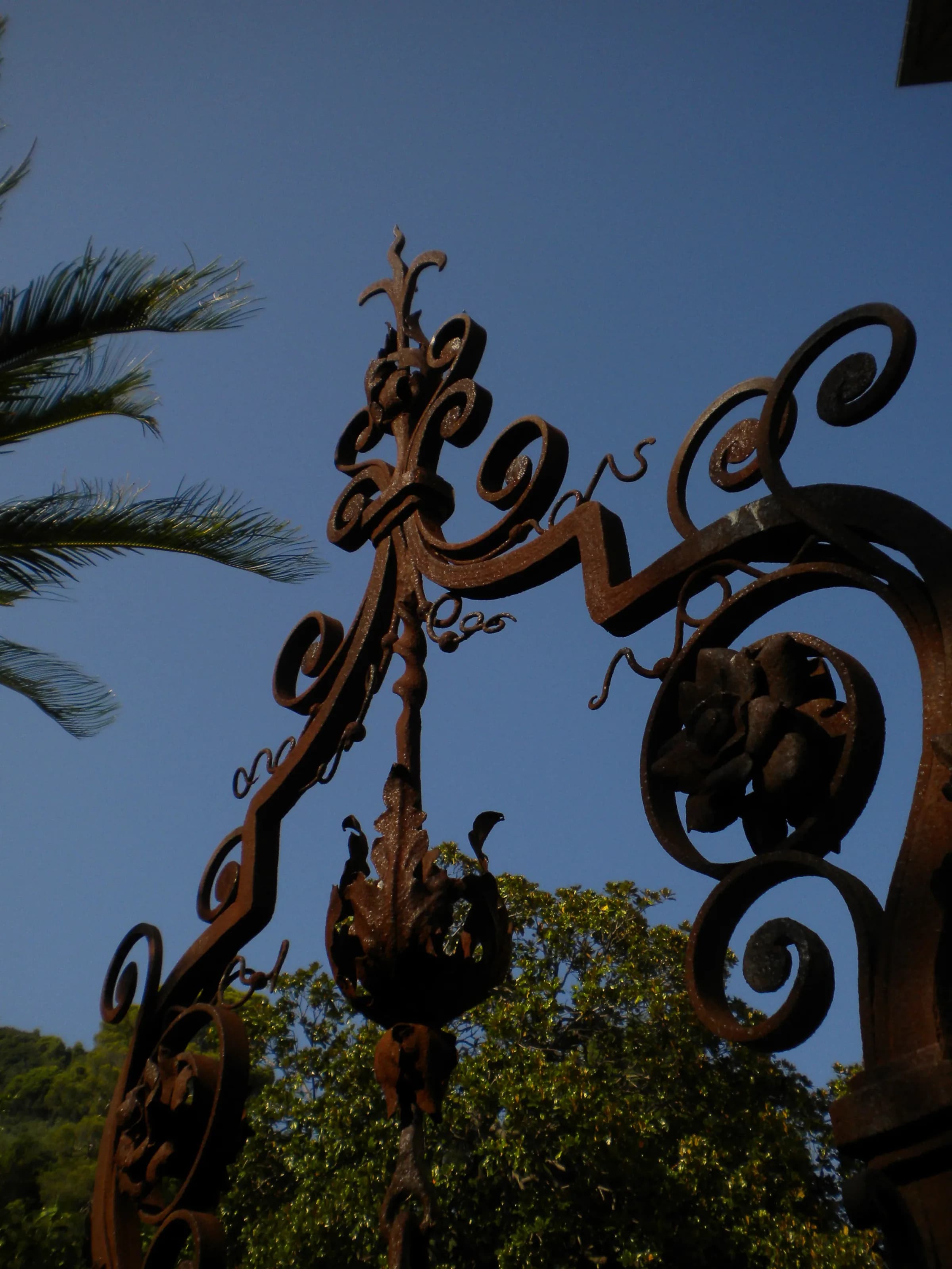 Dettaglio artistico di architettura e design in ferro battuto inquadrato dal basso verso il cielo blu brillante, circondato da alberi e vegetazione rigogliosa in un'atmosfera estiva e mediterranea.